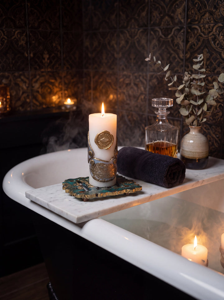 Lit Break the Seal candle with a bronze seal detail on a resin stand across a bathtub, beside a dark towel and glass decanter.