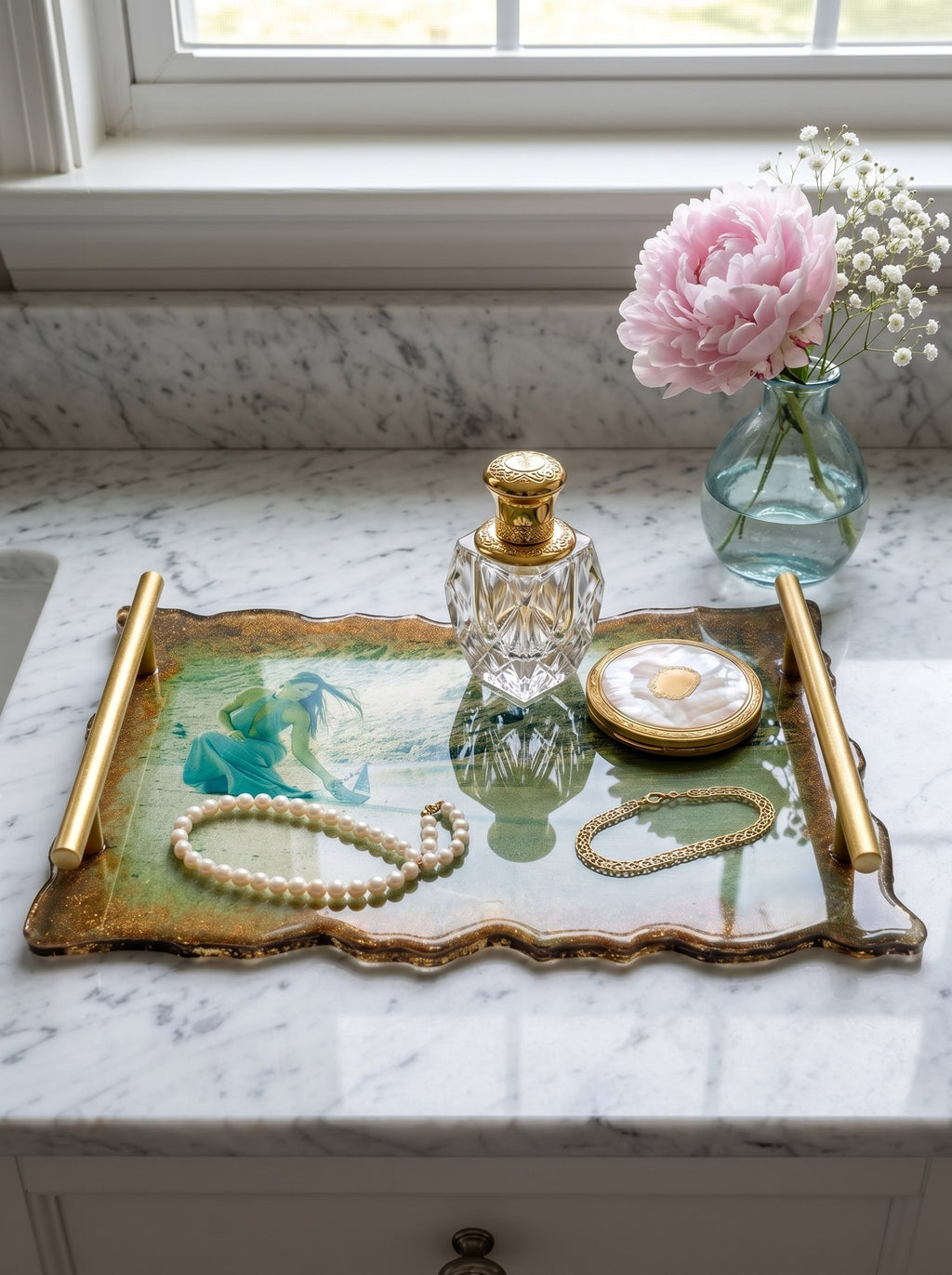 A luxurious handmade resin coffee tray with gold handles resting on a white marble vanity table. Inside the glossy resin, a personalized photo of a woman in a green dress is embedded. The tray is styled with a crystal perfume bottle, a pearl necklace, and soft pink peonies in the background, illuminated by bright natural sunlight.