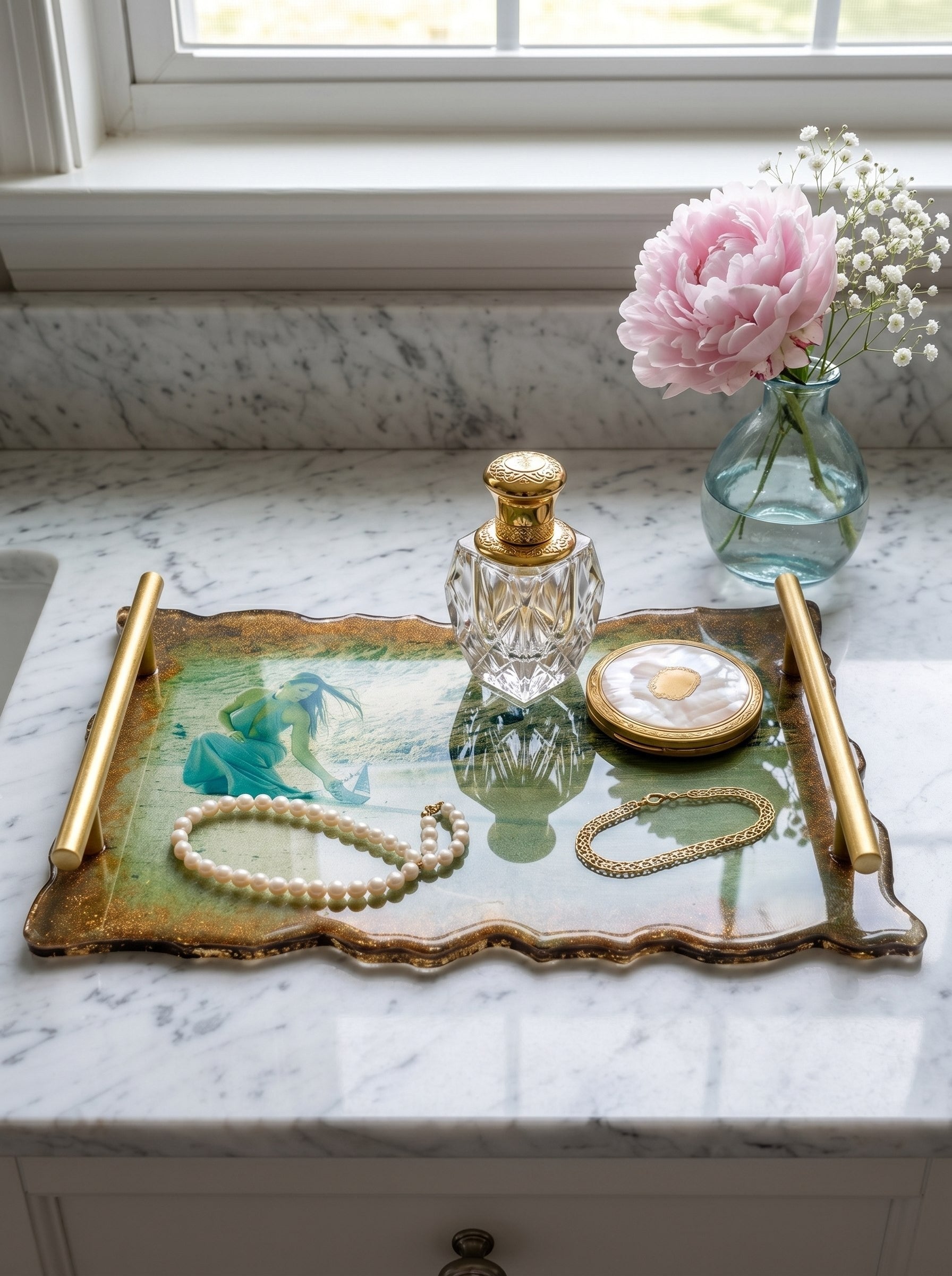 A luxurious handmade resin coffee tray with gold handles resting on a white marble vanity table. Inside the glossy resin, a personalized photo of a woman in a green dress is embedded. The tray is styled with a crystal perfume bottle, a pearl necklace, and soft pink peonies in the background, illuminated by bright natural sunlight.